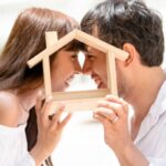 coppia che sorride testa contro testa con in mano una casetta di legno